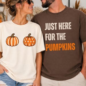 May include: A cream-colored t-shirt featuring two orange pumpkins, one with vertical stripes and one with white polka dots, alongside a dark brown t-shirt with the white and orange text "JUST HERE FOR THE PUMPKINS" printed on the front.