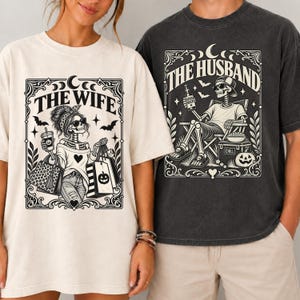 May include: A cream t-shirt featuring a black graphic of a skeleton labeled THE WIFE with shopping bags and a drink, and a charcoal t-shirt featuring a black graphic of a skeleton labeled THE HUSBAND sitting in a chair with a beer and cooler.