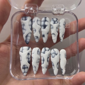 May include: A set of press-on nails in a clear plastic case. The nails are white with a textured, web-like design in shades of gray and black. The nails are almond-shaped.