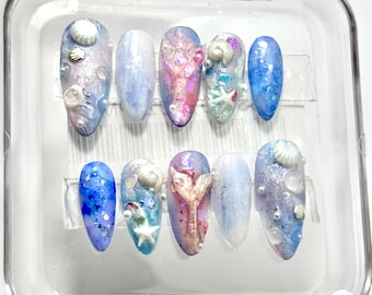 Puede incluir: Un juego de diez uñas artificiales en una caja de plástico transparente. Las uñas presentan una variedad de diseños, incluyendo conchas marinas, purpurina y detalles de perlas. La paleta de colores incluye tonos de azul, rosa y blanco, evocando un tema de playa.