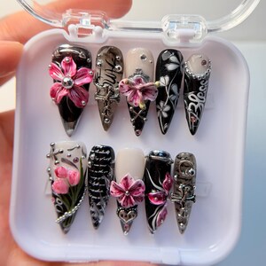 Op de afbeelding: Een set van tien lange, puntige kunstnagels in een doorzichtige plastic doos. De nagels hebben verschillende ontwerpen, waaronder roze bloemen, zilveren accenten en zwart-witte patronen. Sommige nagels hebben het woord "Bless" erop.
