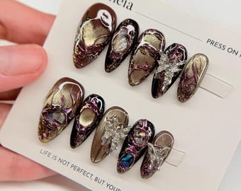 May include: A set of press-on nails with a dark brown, gold, and purple design. Some nails feature silver embellishments. The text "Erytheia" and "Life is not perfect, but your nails are!" are visible.