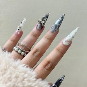 May include: A hand with long, pointed, artificial nails in various designs. One nail is black with a silver accent, others feature marble effects, clear tips with gems, and a silver bow. A pearl ring and a silver ring are also visible.