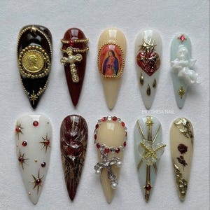 Pode incluir: Uma coleção de dez unhas artificiais longas em forma de amêndoa. Cada unha apresenta designs intrincados, incluindo iconografia religiosa, cruzes e ornamentos dourados e prateados. As cores incluem preto, bordô, creme e ouro.