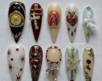 May include: A collection of ten long, almond-shaped artificial nails. Each nail features intricate designs, including religious iconography, crosses, and ornate gold and silver embellishments. Colors include black, burgundy, cream, and gold.