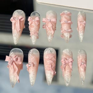 May include: A set of ten long, almond-shaped artificial nails. Each nail features a delicate white lace design and a soft pink bow. Some nails have additional pink embellishments and small pearls.