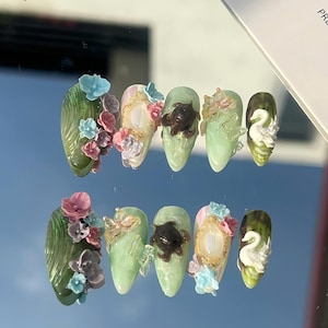 Könnte beinhalten: Ein Set aus zehn mandelförmigen Kunstnägeln. Jeder Nagel weist aufwendige Designs auf, darunter florale Akzente, Miniatur-Schildkröten und Schwanenmotive. Die Farbpalette umfasst Grün-, Rosa- und Blautöne mit goldenen und weißen Details.