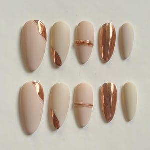 Pode incluir: Um conjunto de dez unhas artificiais em vários designs. As unhas apresentam acabamentos mate e metálicos em tons de nude, pêssego e ouro rosa. Algumas unhas têm um acento metálico em ouro rosa, enquanto outras são de cor sólida.