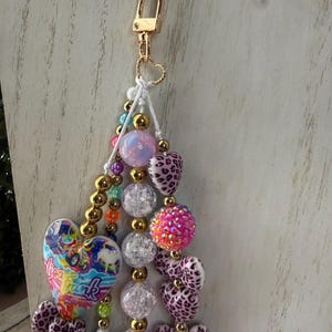 K&ouml;nnte beinhalten: Ein farbenfroher Schl&uuml;sselanh&auml;nger mit einem goldenen Verschluss und einem Herzanh&auml;nger. Der Schl&uuml;sselanh&auml;nger hat eine Vielzahl von Perlen, darunter goldene, klare, rosa und herzf&ouml;rmige Perlen mit Leopardenmuster. Ein herzf&ouml;rmiger Anh&auml;nger mit dem Text "Lisa Frank" ist ebenfalls enthalten.