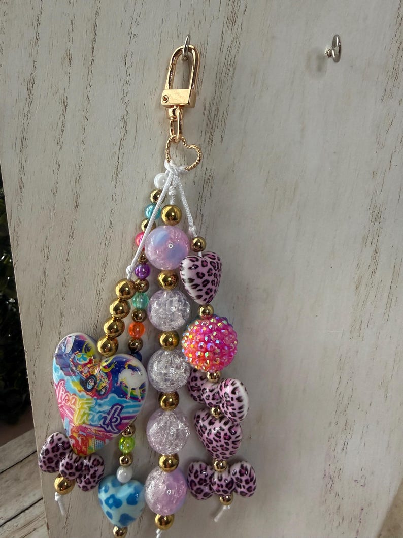 K&ouml;nnte beinhalten: Ein farbenfroher Schl&uuml;sselanh&auml;nger mit einem goldenen Verschluss und einem Herzanh&auml;nger. Der Schl&uuml;sselanh&auml;nger hat eine Vielzahl von Perlen, darunter goldene, klare, rosa und herzf&ouml;rmige Perlen mit Leopardenmuster. Ein herzf&ouml;rmiger Anh&auml;nger mit dem Text "Lisa Frank" ist ebenfalls enthalten.