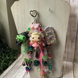 Puede incluir: Un llavero decorativo con dos pequeñas figuras, una verde y otra rubia, unidas a una anilla metálica en forma de corazón. El accesorio incluye varias cuentas de colores, dijes y una etiqueta rectangular con el texto CHANGED FOR GOOD.