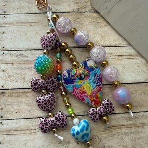 K&ouml;nnte beinhalten: Ein Schl&uuml;sselanh&auml;nger mit Perlen und einem goldfarbenen Verschluss. Der Schl&uuml;sselanh&auml;nger hat eine Vielzahl von Perlen, darunter rosa Herzen und Schleifen mit Leopardenmuster, schillernde Kugeln und einen herzf&ouml;rmigen Anh&auml;nger mit dem Text "Lisa Frank".