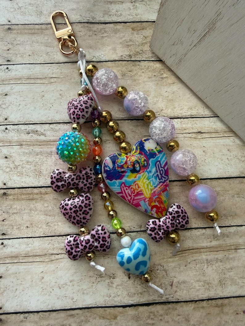 K&ouml;nnte beinhalten: Ein Schl&uuml;sselanh&auml;nger mit Perlen und einem goldfarbenen Verschluss. Der Schl&uuml;sselanh&auml;nger hat eine Vielzahl von Perlen, darunter rosa Herzen und Schleifen mit Leopardenmuster, schillernde Kugeln und einen herzf&ouml;rmigen Anh&auml;nger mit dem Text "Lisa Frank".