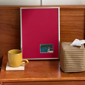 May include: A rectangular silver-framed bulletin board with a solid magenta fabric surface. A small rectangular photograph is attached to the lower right, depicting a person in a red shirt fishing on a wooden dock by a blue lake.