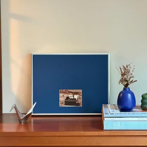 May include: A rectangular picture frame with a thin white border and a deep blue matting, containing a small centered photograph of a person standing near a dark object. The frame is positioned next to a blue vase with dried flowers.