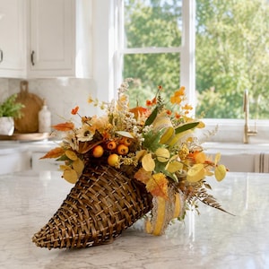 May include: A woven brown cornucopia basket filled with an arrangement of artificial autumn foliage, including orange and yellow leaves, small berries, white flowers, and green sprigs, accented with a decorative yellow and white patterned ribbon.