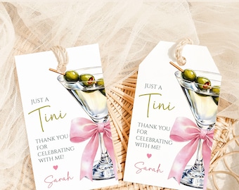 Può includere: Due etichette regalo bianche con un'illustrazione di martini. Ogni etichetta presenta un fiocco rosa, olive e il testo "Just a Tini". Le etichette sono legate con spago e includono il testo "Grazie per aver festeggiato con me!" e un nome.