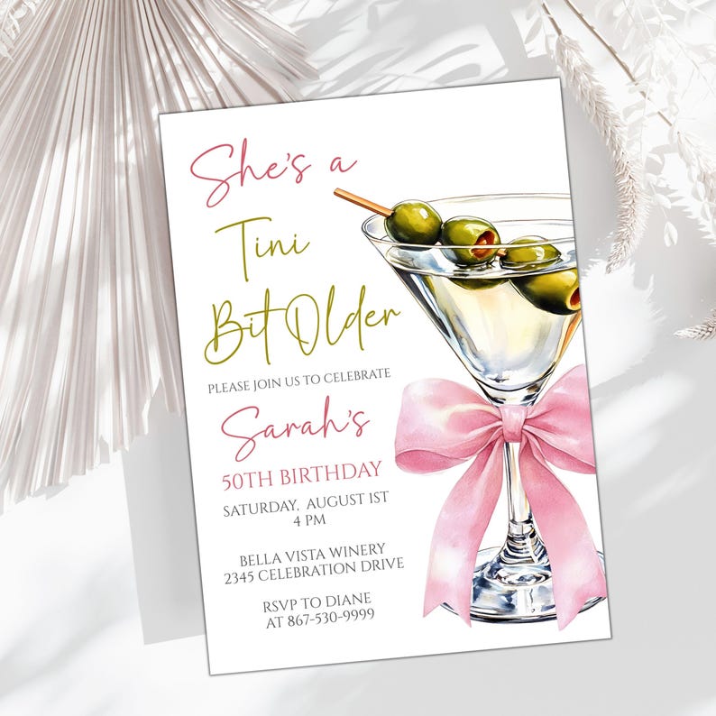 May include: An invitation card with the text "She's a Tini Bit Older" in pink and green, and "Sarah's 50th Birthday". It features a watercolour illustration of a martini glass with olives and a pink bow.