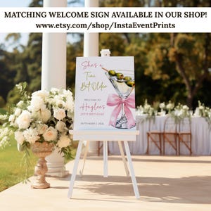 May include: A white easel displays a welcome sign with a martini glass illustration and the text "She's a Tini Bit Older" and "Welcome to Haylee's 25th Birthday September 1, 2026." The sign is decorated with a pink bow and olives. The background includes flowers and a table.