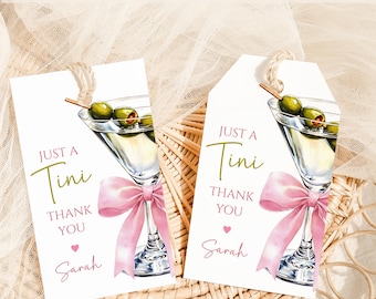 Può includere: Due etichette regalo bianche con un'illustrazione di martini, un fiocco rosa e il testo "Just a Tini, Thank You, Sarah." Le etichette sono appese e su una superficie tessuta.
