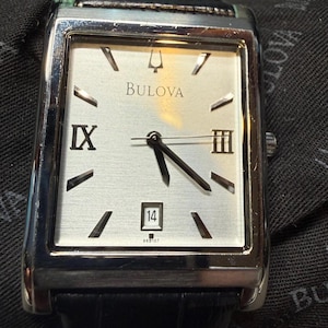 May include: A rectangular Bulova wristwatch with a silver-toned dial, black Roman numerals at the nine and three o'clock positions, silver baton indices, a date window at six o'clock displaying 14, and a black textured leather strap.