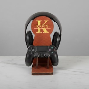 May include: A wooden gaming headset and controller stand. The stand is brown with a gold initial and the name "KEVIN". A black headset and controller are displayed on the stand. The stand is designed to hold gaming accessories.