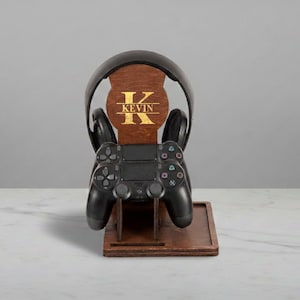 May include: A wooden gaming headset and controller stand. The stand is brown with a gold-colored "K" and the name "KEVIN" engraved on the front. A black headset and controller are placed on the stand.