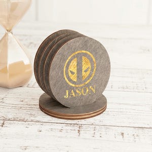May include: A set of gray wooden coasters with a gold Deadpool logo and the name "JASON". The coasters are round and stacked on a matching base. An hourglass is visible in the background.