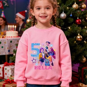 Op de afbeelding: Een roze sweatshirt met een grafisch ontwerp met het nummer "5", sterren en de naam "BELLA". Het ontwerp bevat cartoonfiguren. De sweatshirt wordt gedragen door een jong meisje voor een kerstboom.