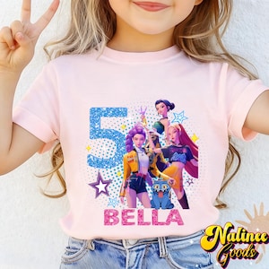 Op de afbeelding: Lichtroze t-shirt met een afbeelding van drie geanimeerde figuren, een blauwe kat, het nummer "5" en de naam "BELLA". Het ontwerp bevat sterren en de woorden "Nalince Goods".