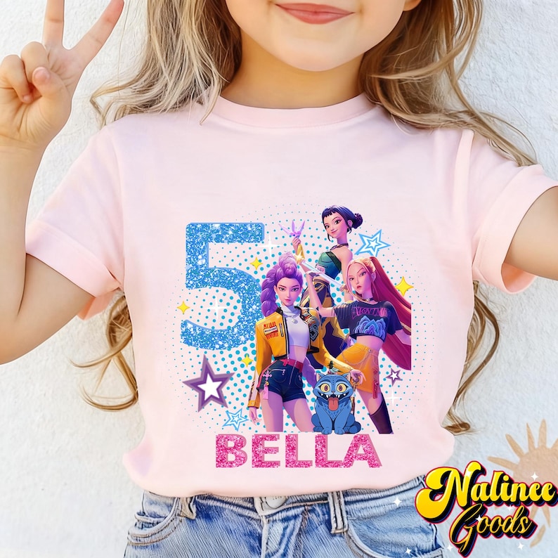 Op de afbeelding: Lichtroze t-shirt met een afbeelding van drie geanimeerde figuren, een blauwe kat, het nummer "5" en de naam "BELLA". Het ontwerp bevat sterren en de woorden "Nalince Goods".