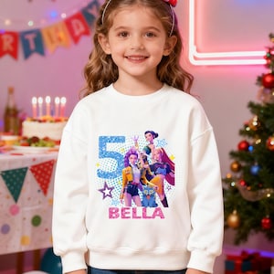 Op de afbeelding: Een witte sweatshirt met een kleurrijke afbeelding met het nummer 5, sterren en de naam "BELLA" in roze. De afbeelding bevat stripfiguren. Het meisje lacht en draagt een haarband.
