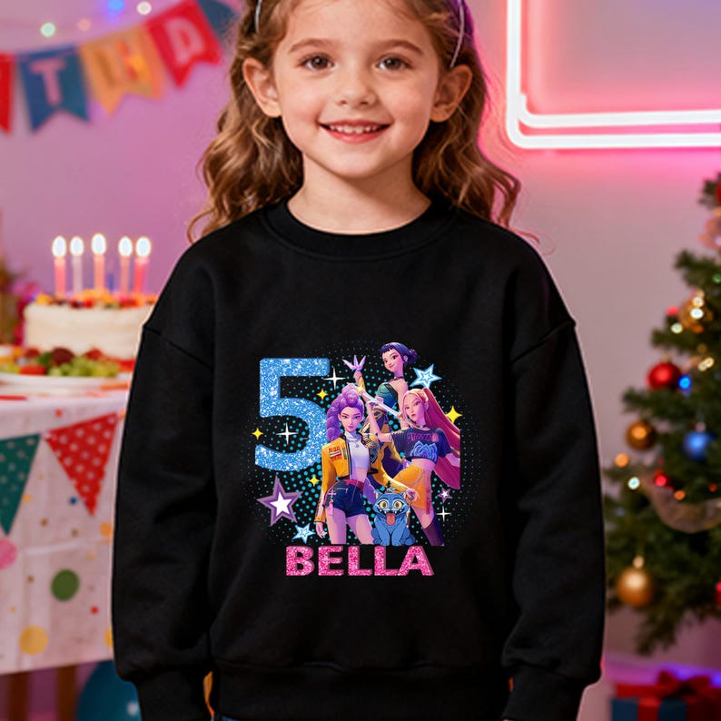 Op de afbeelding: Zwarte sweatshirt met een kleurrijke afbeelding met het nummer 5 in blauw, sterren en de naam "BELLA" in roze. De afbeelding bevat stripfiguren. De sweatshirt wordt door een jong meisje gedragen.