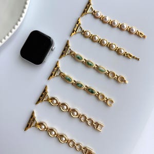 May include: Gold-toned watch bands with pearl and green stone accents. The bands are designed for a smartwatch and feature a variety of decorative links and clasps. A black smartwatch is also visible in the image.