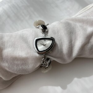 May include: A silver wristwatch with an unusual, asymmetrical face. The watch has a white clock face with black hands and the number 12. The strap includes decorative elements and black and white stones. The watch is displayed on a white surface.