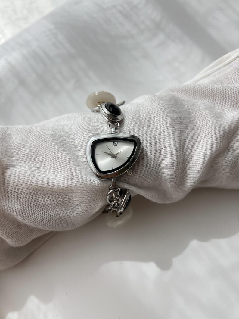 May include: A silver wristwatch with an unusual, asymmetrical face. The watch has a white clock face with black hands and the number 12. The strap includes decorative elements and black and white stones. The watch is displayed on a white surface.