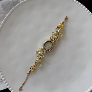 May include: Gold-toned floral bracelet watch with an oval face. The watch features delicate flower and pearl accents along the band. The watch is displayed on a white plate, suggesting a focus on jewelry and accessories.
