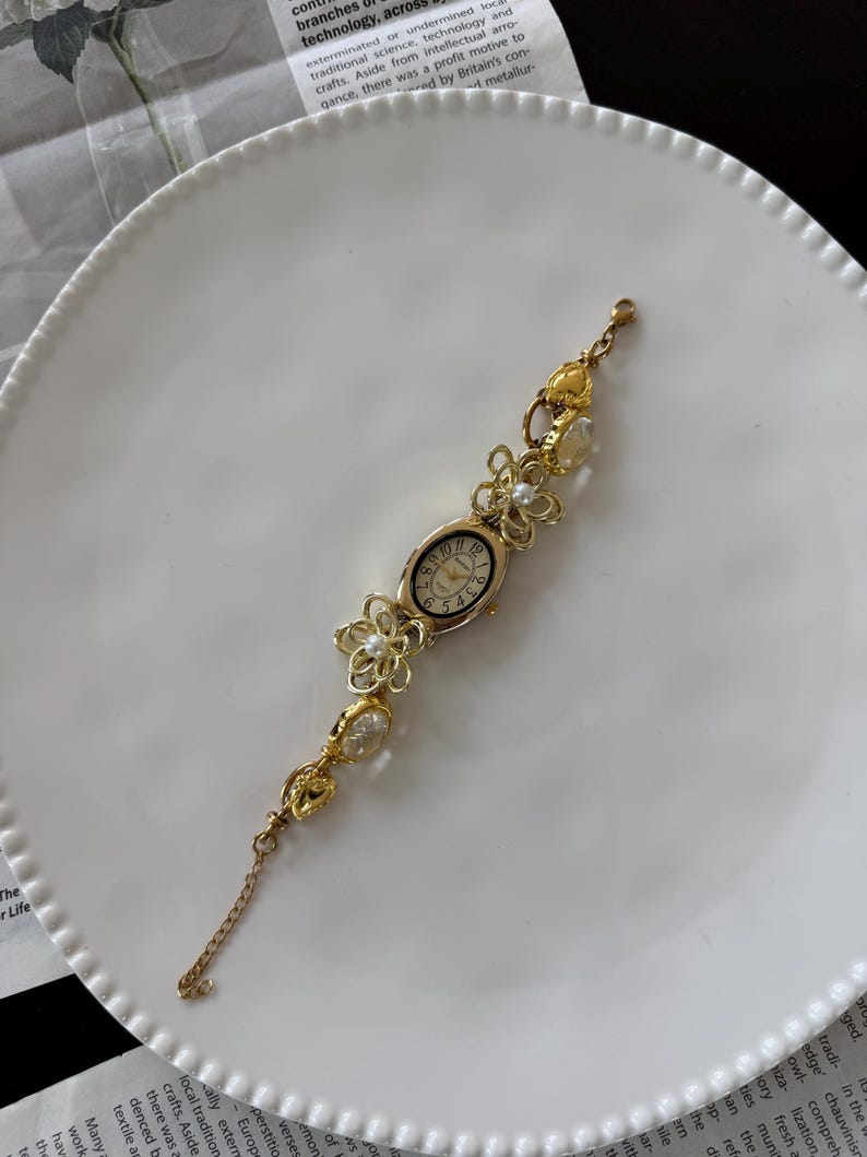 May include: Gold-toned floral bracelet watch with an oval face. The watch features delicate flower and pearl accents along the band. The watch is displayed on a white plate, suggesting a focus on jewelry and accessories.
