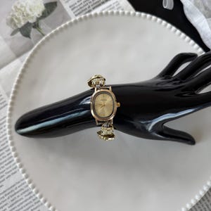 May include: A gold-toned wristwatch with an oval face and a decorative band. The watch is displayed on a black hand-shaped stand, set against a white plate and a backdrop of printed paper.