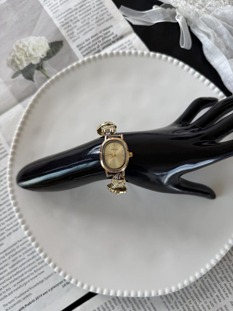 May include: A gold-toned wristwatch with an oval face and a decorative band. The watch is displayed on a black hand-shaped stand, set against a white plate and a backdrop of printed paper.