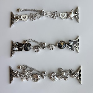 May include: Three silver-toned watch bands featuring decorative charms. The top band has white hearts, flowers, and stars. The middle band includes black circular charms with gold flecks and butterfly shapes. The bottom band features textured flowers and pearls.