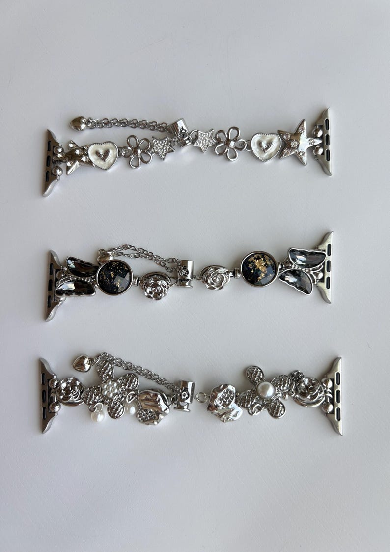 May include: Three silver-toned watch bands featuring decorative charms. The top band has white hearts, flowers, and stars. The middle band includes black circular charms with gold flecks and butterfly shapes. The bottom band features textured flowers and pearls.