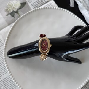 May include: A gold-toned wristwatch with a burgundy oval face and a burgundy rose accent. The watch is on a black hand-shaped display, set against a white plate and newspaper background. The watch face reads "NAIDU".