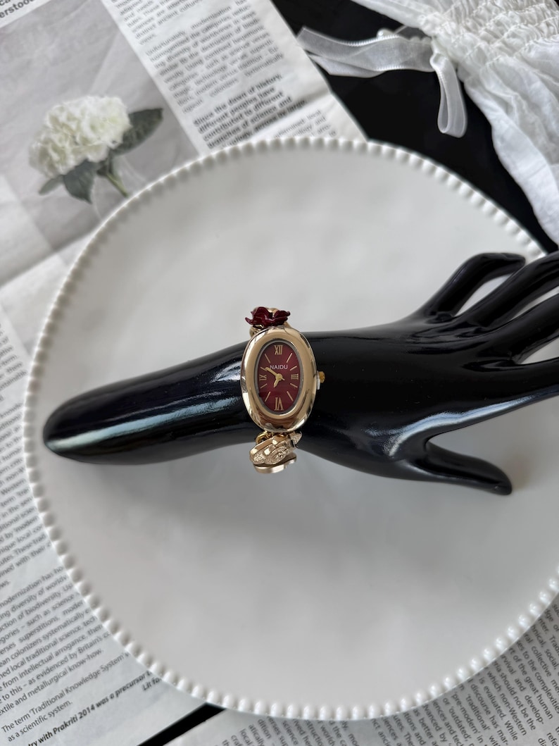 May include: A gold-toned wristwatch with a burgundy oval face and a burgundy rose accent. The watch is on a black hand-shaped display, set against a white plate and newspaper background. The watch face reads "NAIDU".