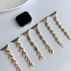 May include: Gold-toned watch bands with various designs, including faux pearls and green oval-shaped stones. A black smartwatch is also present. The bands are displayed on a white surface, showcasing their elegant and decorative appeal.