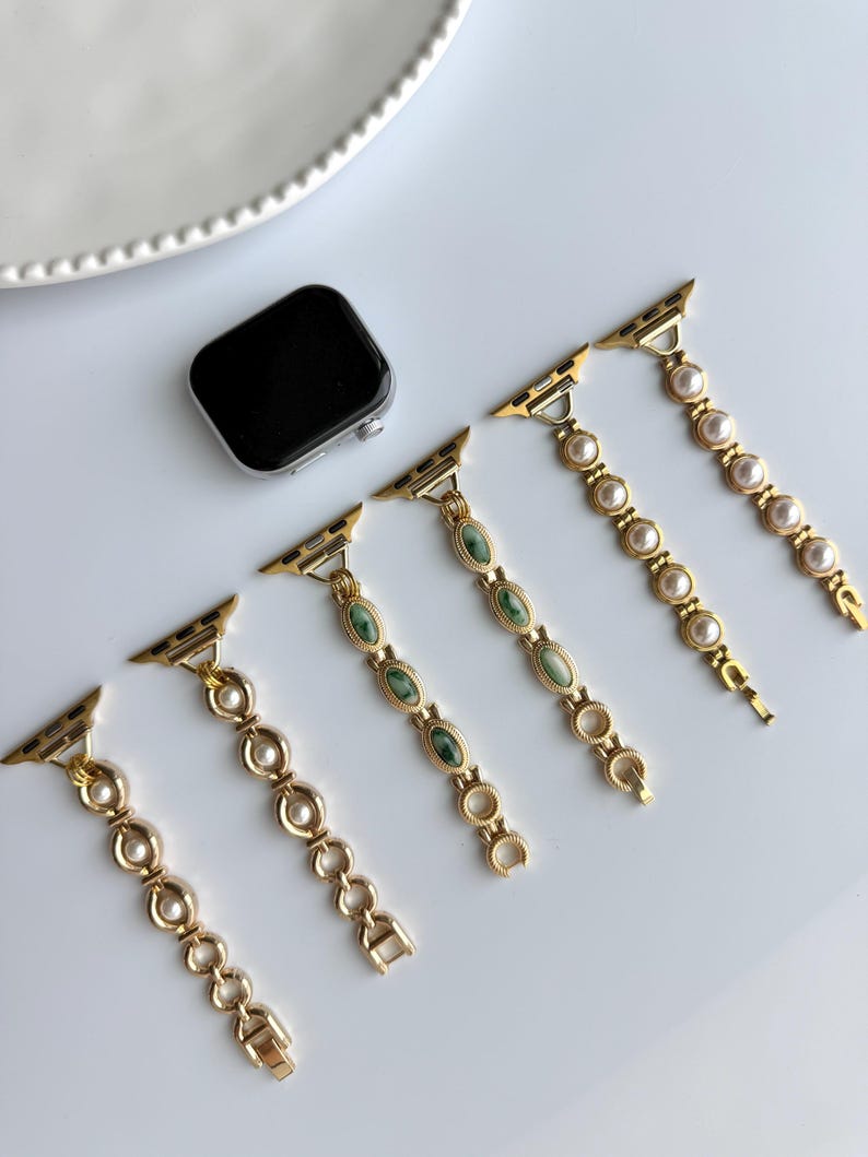 May include: Gold-toned watch bands with various designs, including faux pearls and green oval-shaped stones. A black smartwatch is also present. The bands are displayed on a white surface, showcasing their elegant and decorative appeal.