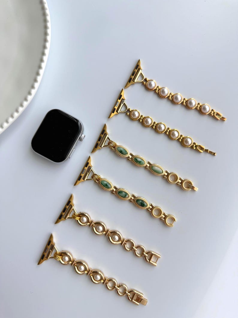 May include: Gold-toned watch bands with pearl and green stone accents. The bands are designed for a smartwatch and feature a variety of decorative links and clasps. A black smartwatch is also visible in the image.