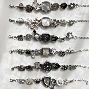 May include: Six silver bracelet watches with decorative charms such as hearts, stars, flowers, and geometric shapes. Each watch has a different face, with square and round dials. The bracelets are displayed on a white surface.