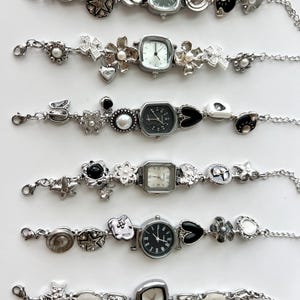 May include: Six silver charm bracelets with watch faces. Each bracelet has a different watch face shape, surrounded by decorative charms such as hearts, stars, flowers, and pearls. The watch faces have black or white dials.