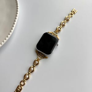 May include: A gold-toned watch band with a pearl accent is attached to a silver smartwatch with a black screen. The band has a chain-like design with oval-shaped pearls. The watch and band are on a white surface.
